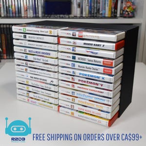 May include: A black game cartridge holder filled with 3DS video games. Titles include "Animal Crossing: New Leaf", "Mario Kart 7", and several Pokémon games. The holder is on a white surface.