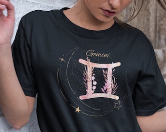 Puede incluir: Camiseta negra con un diseño floral rosa y dorado del símbolo del zodíaco Géminis. El texto "Géminis" está escrito en dorado sobre el símbolo.