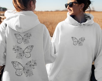 May include: Two white hoodies with a black butterfly and floral design. The hoodie on the left has three butterflies and the hoodie on the right has one butterfly.