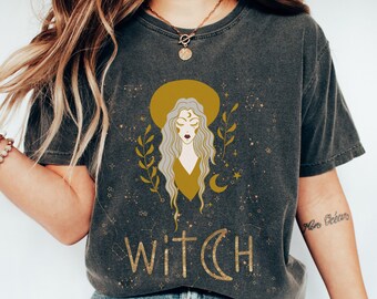 Könnte beinhalten: Ein dunkelgraues T-Shirt mit einem Motiv einer Frau mit langen blonden Haaren, die eine gelbe Krone und einen Halbmond trägt. Der Text "WITCH" ist unter dem Motiv gedruckt.