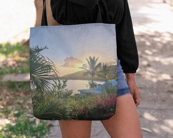 Puede incluir: Una bolsa de tela con una puesta de sol sobre una isla tropical. La bolsa está hecha de lona y tiene una correa larga. La escena presenta palmeras, una masa de agua y un pequeño edificio.