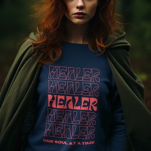 Könnte beinhalten: Ein marineblaues Sweatshirt mit dem Wort "HEALER" fünfmal in einem Retro-Schriftstil gestapelt, wobei das mittlere Wort rot ist. Der Satz "ONE SOUL AT A TIME" ist darunter gedruckt. Die Person trägt einen grünen Kapuzenmantel.