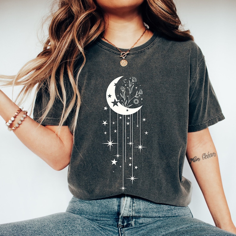 May include: A dark grey t-shirt featuring a white crescent moon design. The moon has a floral design inside and stars hanging from it. The shirt has a relaxed fit and short sleeves.