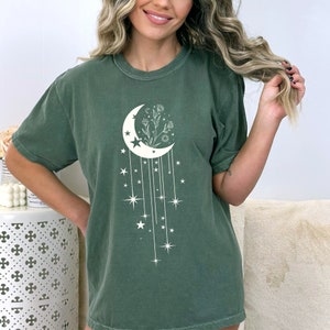May include: A green t-shirt featuring a white crescent moon design. The moon is surrounded by flowers and stars. The stars are connected to the moon by lines.