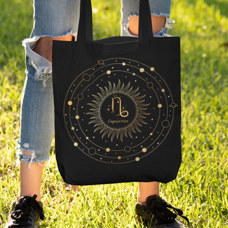 May include: Black tote bag with a gold sun and zodiac symbol for Capricorn. The sun has a circle around it with stars and planets.