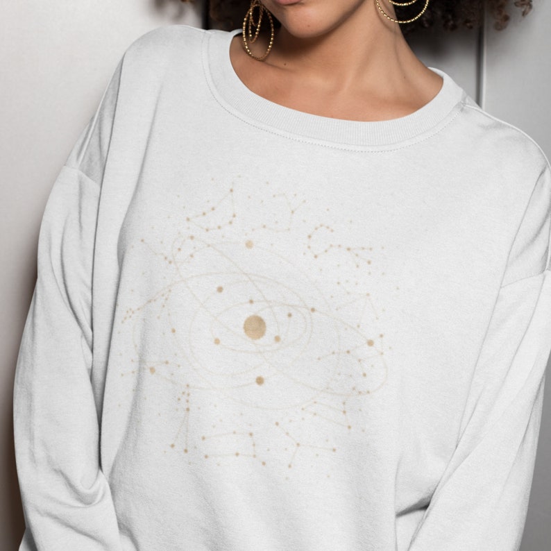 Puede incluir: Sudadera blanca con un dise&ntilde;o de constelaci&oacute;n dorada en la parte delantera. El dise&ntilde;o presenta un c&iacute;rculo grande con c&iacute;rculos m&aacute;s peque&ntilde;os y l&iacute;neas que irradian hacia afuera.