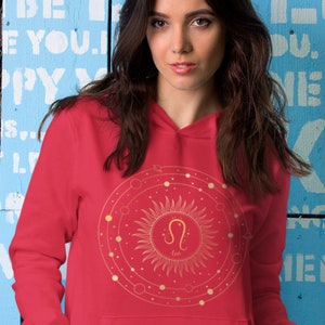 May include: Red hooded sweatshirt featuring a gold zodiac symbol of Leo, a lion, within a circle containing a sun and stars. The text "Leo" is positioned beneath the symbol.