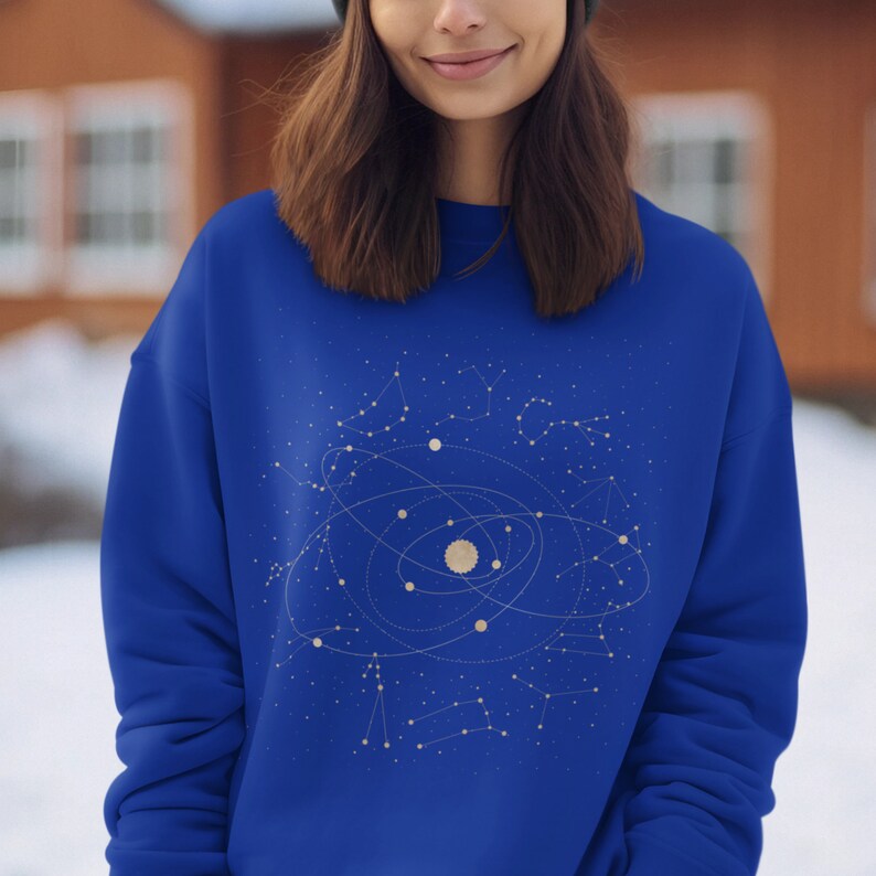 Puede incluir: Una sudadera azul real con un dise&ntilde;o de constelaci&oacute;n dorada que representa una galaxia y planetas. El dise&ntilde;o est&aacute; centrado en la parte delantera de la sudadera.