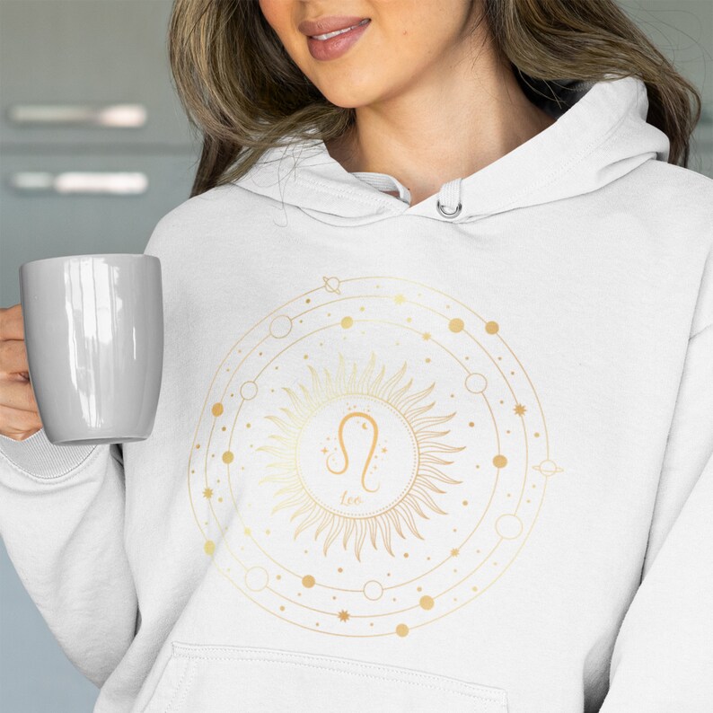 May include: White hoodie featuring a gold zodiac symbol for Leo, encircled by a sun and celestial design.