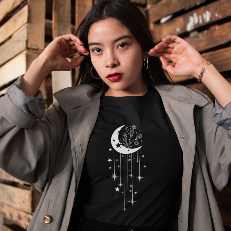 May include: A black t-shirt featuring a white crescent moon design. The moon is filled with a floral pattern and has stars and dangling stars hanging from it.