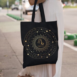 May include: Black tote bag with a gold sun and zodiac symbol for Capricorn. The sun has a circle around it with planets and stars.
