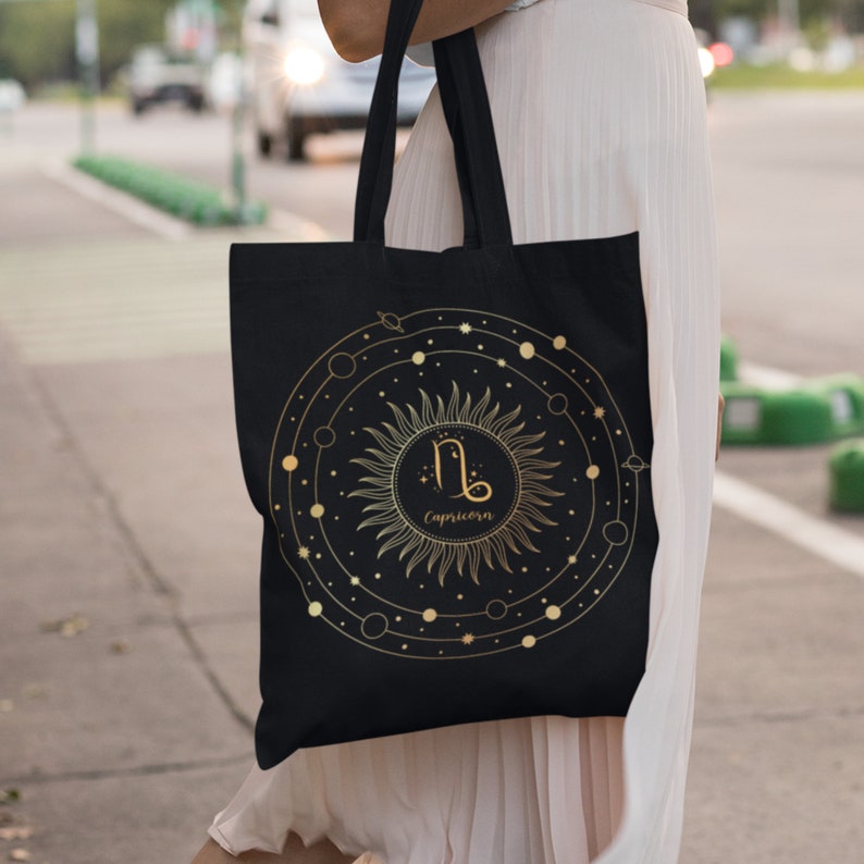 May include: Black tote bag with a gold sun and zodiac symbol for Capricorn. The sun has a circle around it with planets and stars.