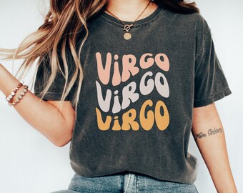 Może przedstawiać: Szary t-shirt z napisem "Virgo" powtórzonym trzy razy w retro czcionce. Słowo "Virgo" jest w kolorze różowym, szarym i żółtym.