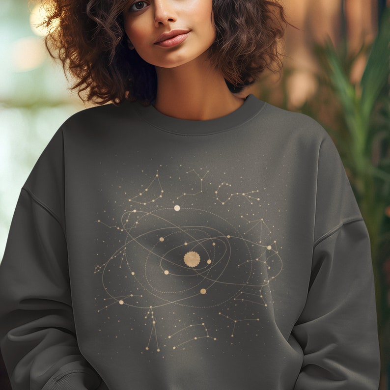 Puede incluir: Una sudadera gris oscuro con un gr&aacute;fico de un sistema solar con constelaciones y planetas en color beige claro.
