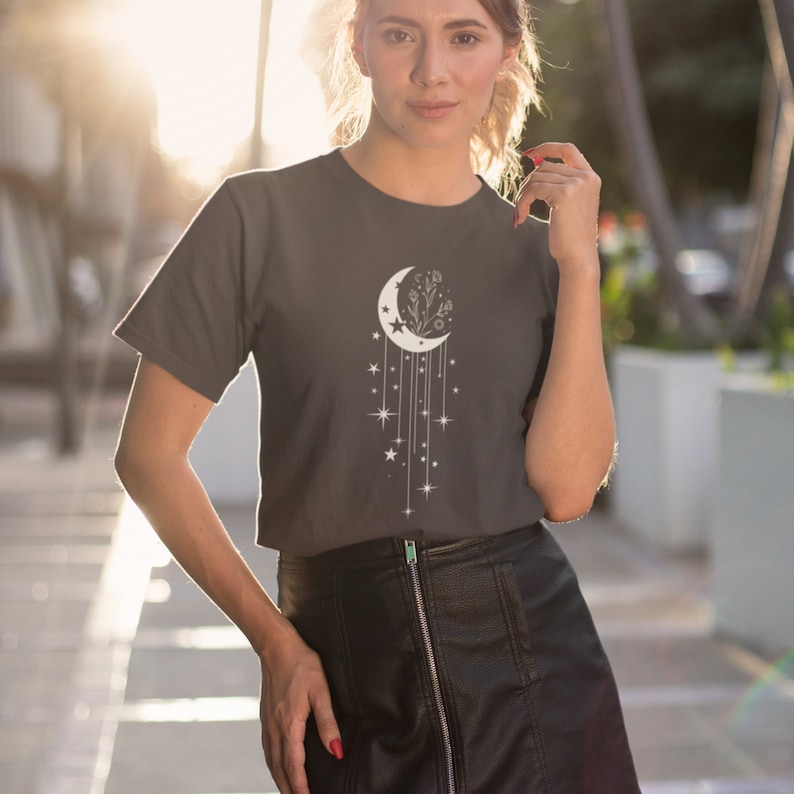 May include: A dark grey t-shirt with a white crescent moon design. The moon has a floral design inside and is surrounded by stars and dangling lines.