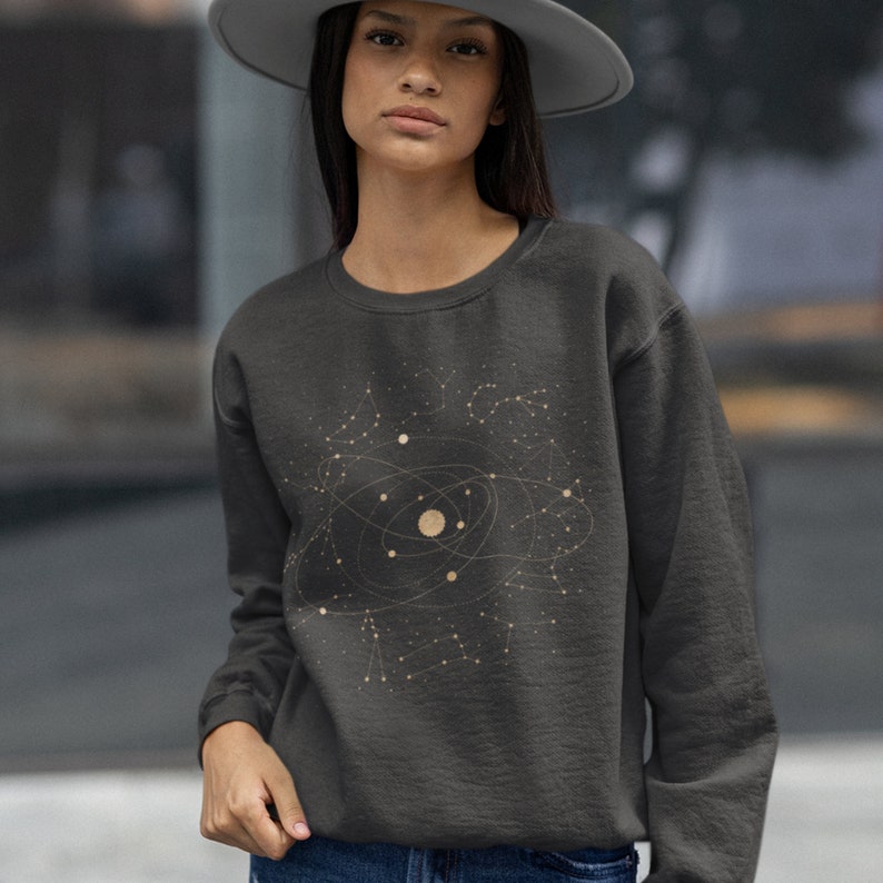 Puede incluir: Una sudadera gris oscuro con un gr&aacute;fico dorado de un sistema solar y constelaciones. El gr&aacute;fico incluye un c&iacute;rculo grande con c&iacute;rculos m&aacute;s peque&ntilde;os dentro, que representan planetas, y una constelaci&oacute;n de estrellas.