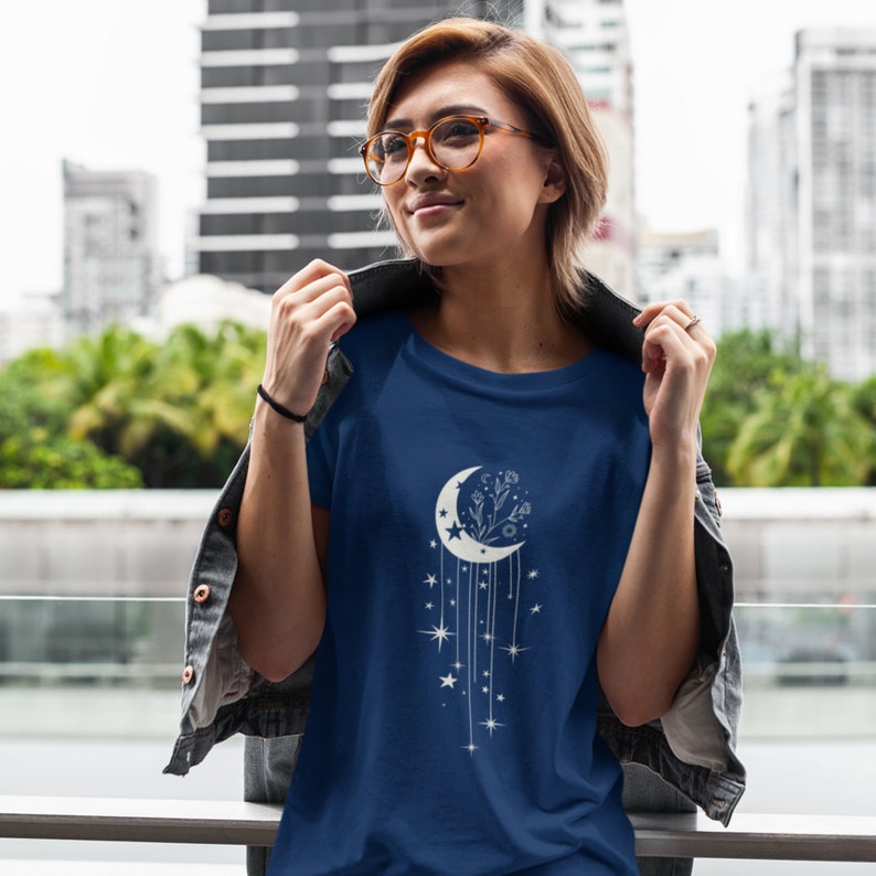 May include: A navy blue t-shirt featuring a white crescent moon design. The moon is decorated with a floral motif and dangling stars.