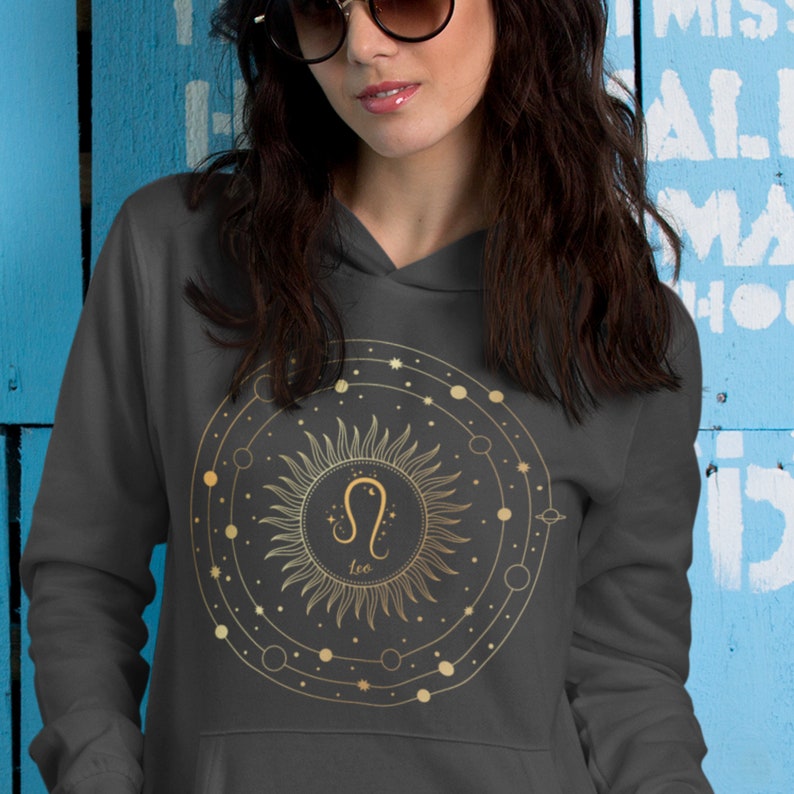 May include: A grey hoodie featuring a gold sun and Leo zodiac symbol design. The sun is surrounded by a circle with small stars and planets.