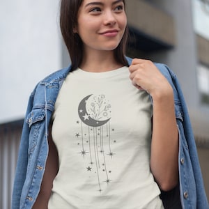 May include: A white t-shirt featuring a grey crescent moon design. The moon is filled with a floral design and has stars hanging from it.