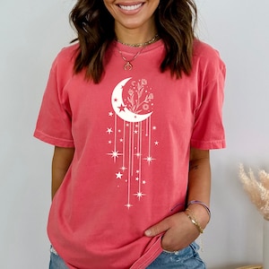 May include: A coral pink t-shirt featuring a white crescent moon design. The moon has a floral design inside and is surrounded by stars.