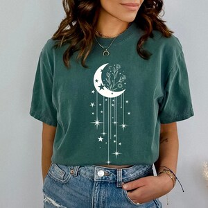 May include: A teal green t-shirt featuring a white crescent moon design. The moon has a floral design inside and is surrounded by stars and dangling lines.