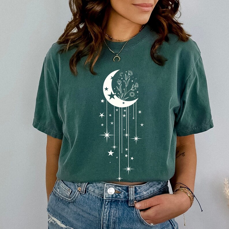 May include: A teal green t-shirt featuring a white crescent moon design. The moon has a floral design inside and is surrounded by stars and dangling lines.