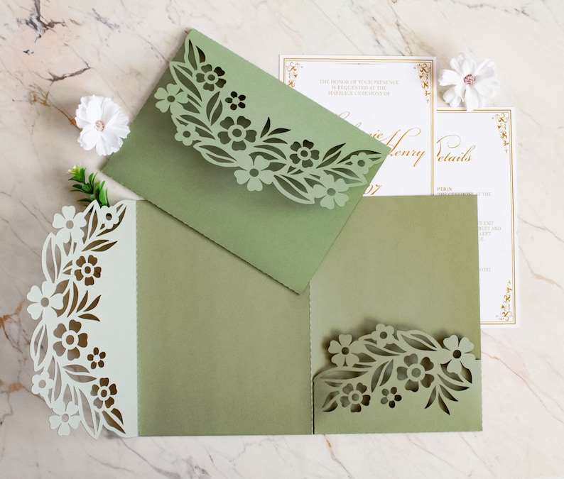 May include: Green laser cut wedding invitation with floral design. The invitation is folded in thirds and has a white inner layer. The text on the invitation reads "The honor of your presence is requested at the marriage ceremony of".