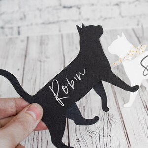 K&ouml;nnte beinhalten: Zwei Papier-Katzen-Platzkartehalter, einer schwarz und einer wei&szlig;. Die schwarze Katze hat den Namen "Robin" in wei&szlig; darauf geschrieben. Die wei&szlig;e Katze hat den Namen "Sand" in schwarz darauf geschrieben.