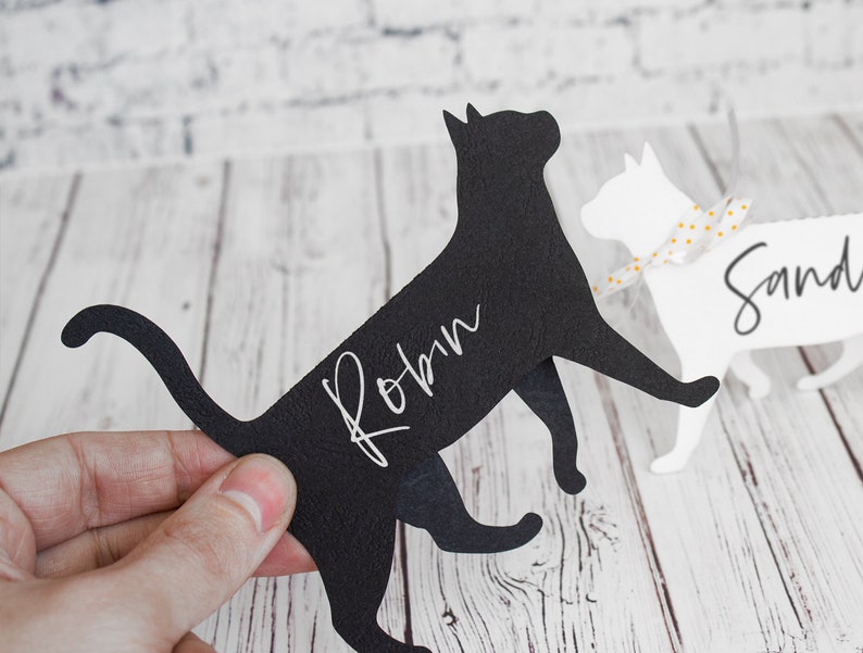 K&ouml;nnte beinhalten: Zwei Papier-Katzen-Platzkartehalter, einer schwarz und einer wei&szlig;. Die schwarze Katze hat den Namen "Robin" in wei&szlig; darauf geschrieben. Die wei&szlig;e Katze hat den Namen "Sand" in schwarz darauf geschrieben.