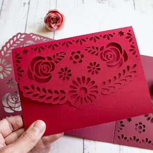 Puede incluir: Una tarjeta de papel roja con un dise&ntilde;o decorativo de corte con flores y hojas. La tarjeta est&aacute; doblada por la mitad y tiene un borde festoneado.