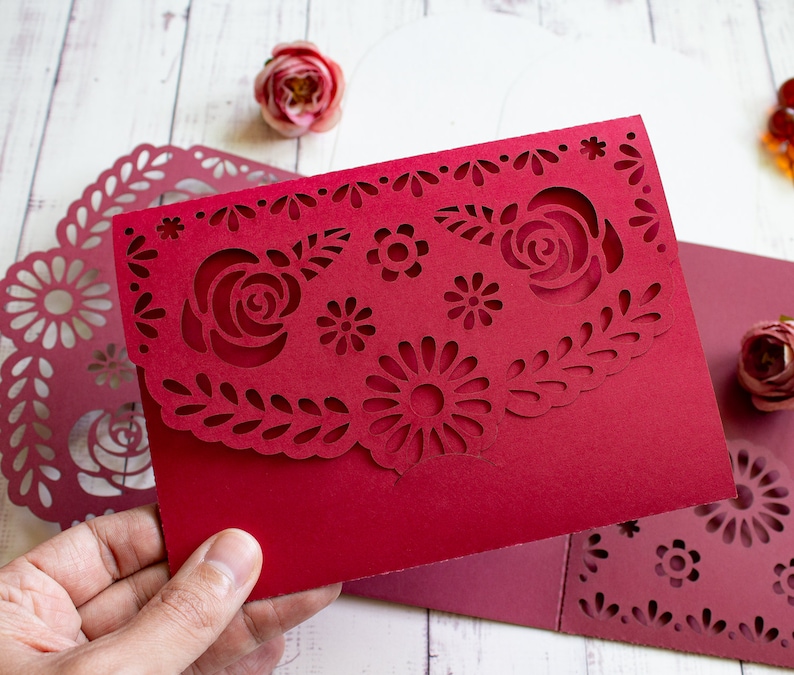 Puede incluir: Una tarjeta de papel roja con un dise&ntilde;o decorativo de corte con flores y hojas. La tarjeta est&aacute; doblada por la mitad y tiene un borde festoneado.