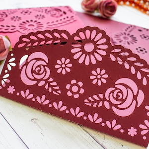Puede incluir: Una tarjeta de papel burdeos con un dise&ntilde;o floral rosa. La tarjeta tiene un borde festoneado y est&aacute; decorada con flores y hojas recortadas.