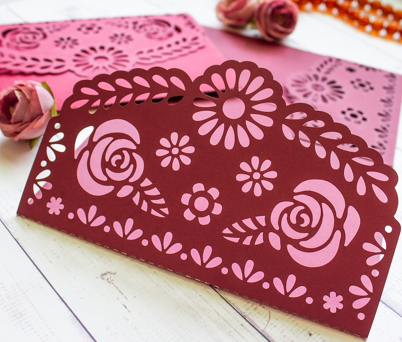 Puede incluir: Una tarjeta de papel burdeos con un dise&ntilde;o floral rosa. La tarjeta tiene un borde festoneado y est&aacute; decorada con flores y hojas recortadas.
