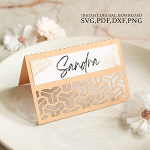 Può includere: Un segnaposto beige con un motivo geometrico e un'etichetta bianca con il nome "Sandra" scritto in calligrafia nera. L'etichetta ha un design geometrico dorato sullo sfondo.