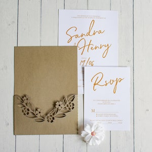 May include: A brown envelope with a laser-cut floral design. The envelope is open to reveal a white card with the text "Sandra & Henry 19/06" in gold script. A second white card with the text "RSVP" in gold script is also visible.