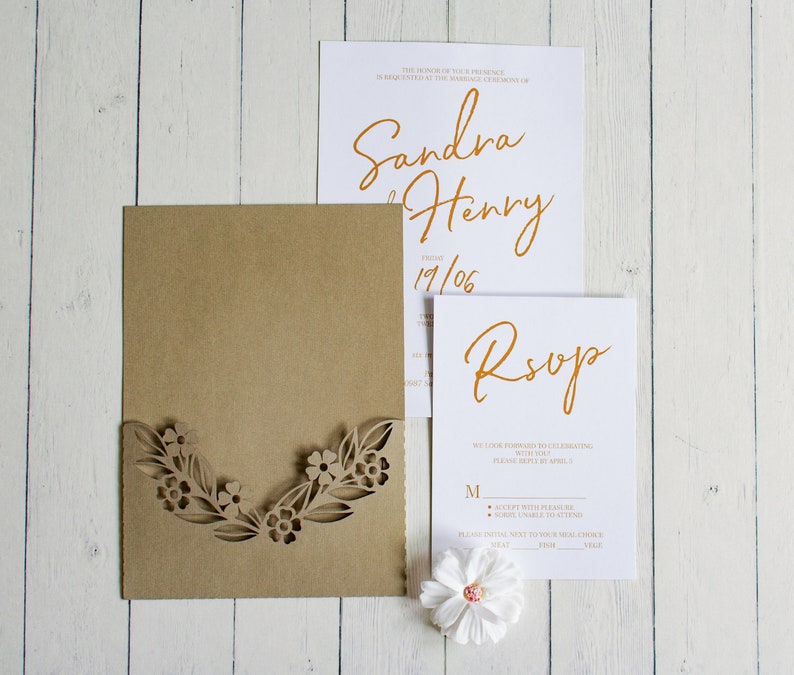 May include: A brown envelope with a laser-cut floral design. The envelope is open to reveal a white card with the text "Sandra & Henry 19/06" in gold script. A second white card with the text "RSVP" in gold script is also visible.