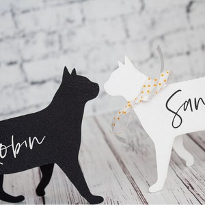 K&ouml;nnte beinhalten: Zwei schwarze und wei&szlig;e Katzen-Silhouetten-Platzkarte mit den Namen "Robin" und "Sandra" in wei&szlig;. Die wei&szlig;e Katze hat ein wei&szlig;es und gelbes gepunktetes Band um den Hals.