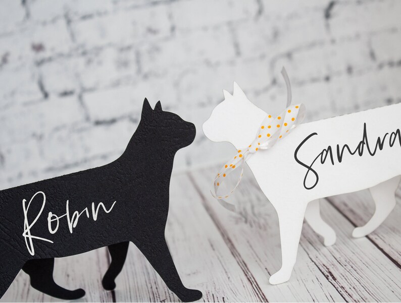 K&ouml;nnte beinhalten: Zwei schwarze und wei&szlig;e Katzen-Silhouetten-Platzkarte mit den Namen "Robin" und "Sandra" in wei&szlig;. Die wei&szlig;e Katze hat ein wei&szlig;es und gelbes gepunktetes Band um den Hals.