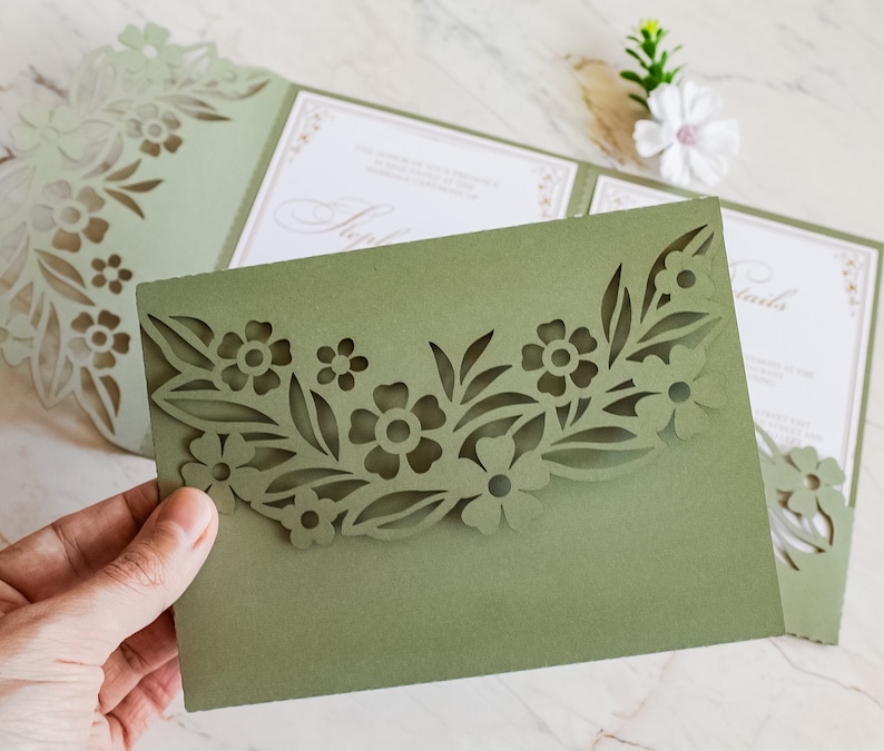 May include: A green folded card with a cut-out design of flowers and leaves. The card is folded in half and the front flap has a cut-out design of flowers and leaves. The card is blank inside.