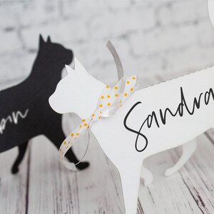 K&ouml;nnte beinhalten: Zwei schwarze und wei&szlig;e katzenf&ouml;rmige Platzkartehalter. Die wei&szlig;e Katze hat ein gelbes und wei&szlig;es gepunktetes Band um den Hals gebunden und den Namen "Sandra" darauf geschrieben. Die schwarze Katze hat den Namen "Robin" darauf geschrieben.