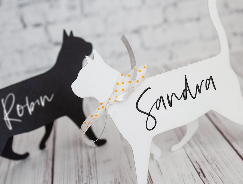 K&ouml;nnte beinhalten: Zwei schwarze und wei&szlig;e katzenf&ouml;rmige Platzkartehalter. Die wei&szlig;e Katze hat ein gelbes und wei&szlig;es gepunktetes Band um den Hals gebunden und den Namen "Sandra" darauf geschrieben. Die schwarze Katze hat den Namen "Robin" darauf geschrieben.