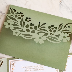 May include: Sage green wedding invitation envelope with a laser cut floral design. The text on the invitation reads "The honor of your presence is requested at the marriage ceremony of"