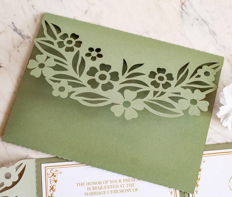 May include: Sage green wedding invitation envelope with a laser cut floral design. The text on the invitation reads "The honor of your presence is requested at the marriage ceremony of"