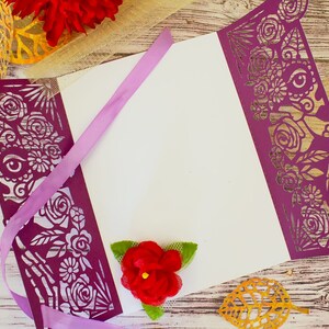 May include: Purple laser cut paper with a floral and skeletal design. The paper is folded to create a card or invitation. A purple ribbon is tied around the card. A red flower is on the card.