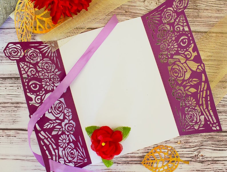 May include: Purple laser cut paper with a floral and skeletal design. The paper is folded to create a card or invitation. A purple ribbon is tied around the card. A red flower is on the card.