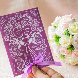 May include: A purple paper cut-out wedding invitation with a floral design and a skull with eyes. The invitation is tied with a purple ribbon and is being held by a hand.