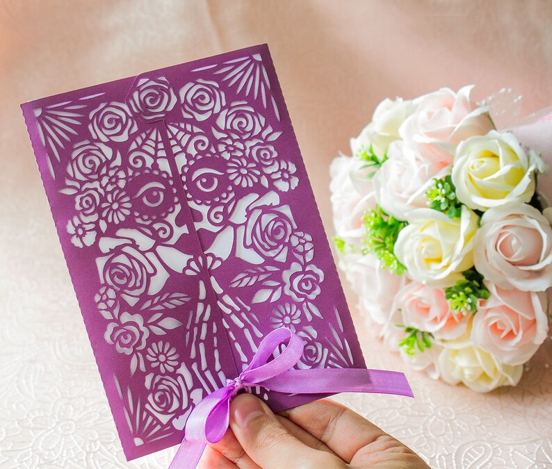 May include: A purple paper cut-out wedding invitation with a floral design and a skull with eyes. The invitation is tied with a purple ribbon and is being held by a hand.