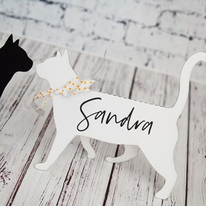K&ouml;nnte beinhalten: Wei&szlig;er Papierschnitt eines Katers mit einem gelben und wei&szlig;en gepunkteten Band um den Hals. Die Katze steht auf einer wei&szlig;en Holzoberfl&auml;che. Der Name "Sandra" ist auf den K&ouml;rper der Katze geschrieben.