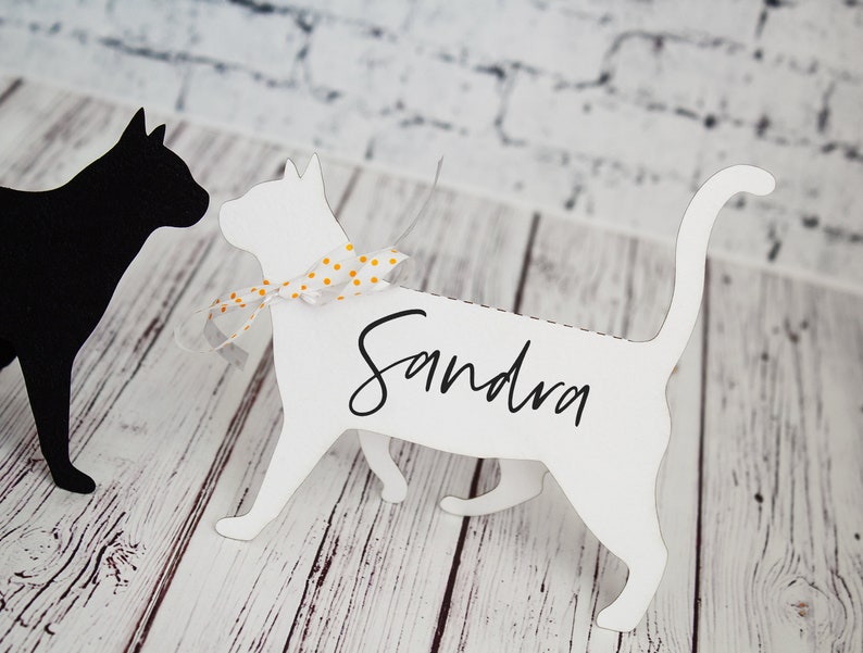 K&ouml;nnte beinhalten: Wei&szlig;er Papierschnitt eines Katers mit einem gelben und wei&szlig;en gepunkteten Band um den Hals. Die Katze steht auf einer wei&szlig;en Holzoberfl&auml;che. Der Name "Sandra" ist auf den K&ouml;rper der Katze geschrieben.