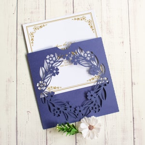 May include: A purple invitation card with a laser cut floral design. The card is folded in half and the front has a white insert with gold lettering that says "Save the Date".